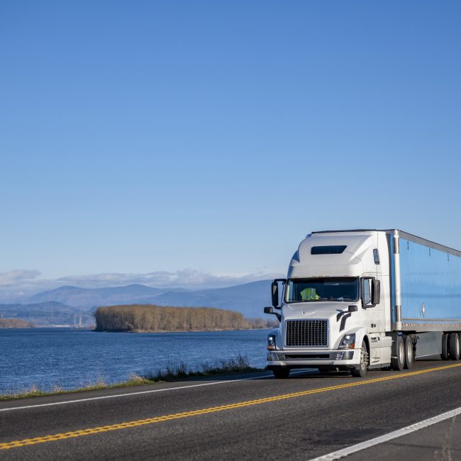 Commercial big rig classic white industrial grade long haul professional semi truck transporting cargo in dry van semi trailer moving on the narrow winding road along the Columbia River
